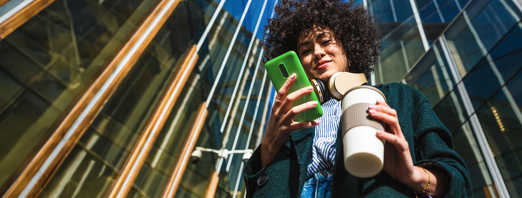 Woman using her mobile phone holding cup of coffee in front of building